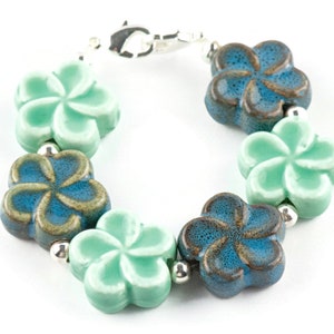 May include: A bracelet featuring alternating blue and green ceramic flower charms. The flowers have a textured finish and are linked by silver beads.