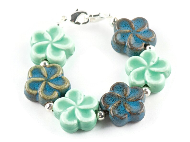 May include: A bracelet featuring alternating blue and green ceramic flower charms. The flowers have a textured finish and are linked by silver beads.