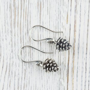May include: A pair of silver earrings with a pine cone design. The earrings are on a white wooden surface.