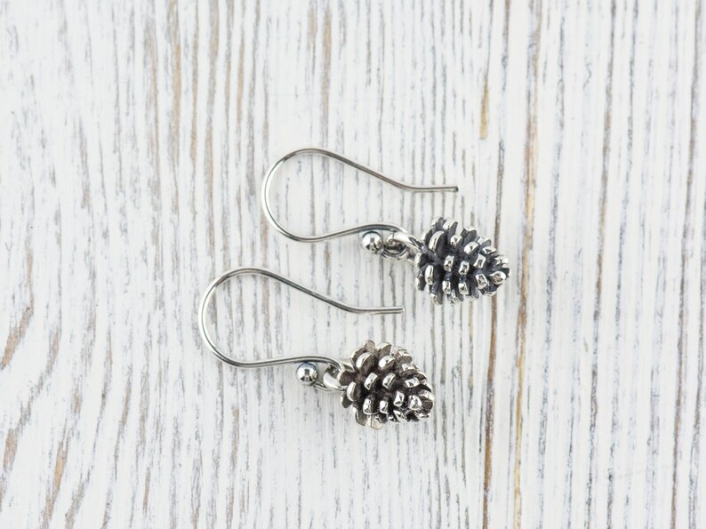 May include: A pair of silver earrings with a pine cone design. The earrings are on a white wooden surface.