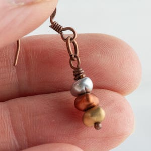 May include: A handcrafted earring with a copper-colored hook and chain. The earring features three beads in silver, copper, and gold colors. The beads are strung together, creating a simple yet elegant design.