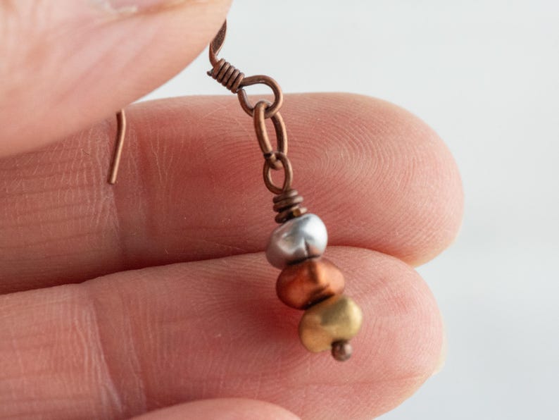 May include: A handcrafted earring with a copper-colored hook and chain. The earring features three beads in silver, copper, and gold colors. The beads are strung together, creating a simple yet elegant design.
