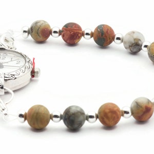 May include: A silver Geneva watch with a white face and a beaded bracelet. The bracelet features multi-colored, round gemstone beads and small silver beads. The watch has a decorative bezel and a red crown.