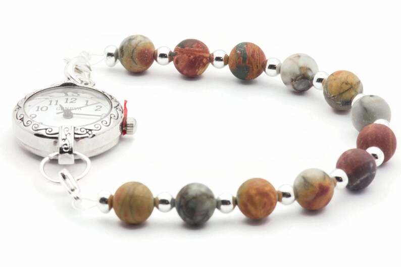 May include: A silver Geneva watch with a white face and a beaded bracelet. The bracelet features multi-colored, round gemstone beads and small silver beads. The watch has a decorative bezel and a red crown.