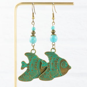 K&ouml;nnte beinhalten: Ein Paar Ohrringe mit Anh&auml;nger, die t&uuml;rkisfarbene Perlen und bronzefarbene Fisch-Anh&auml;nger mit aufwendigen gr&uuml;nen Designs zeigen.