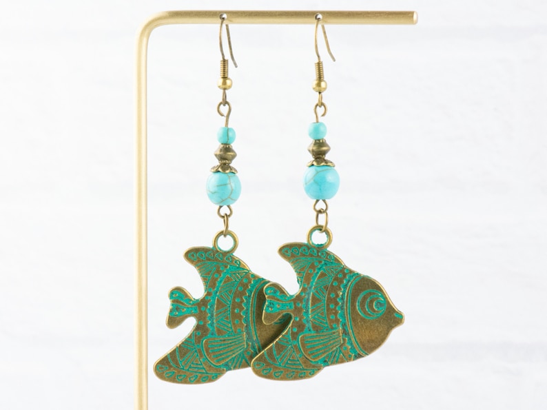 K&ouml;nnte beinhalten: Ein Paar Ohrringe mit Anh&auml;nger, die t&uuml;rkisfarbene Perlen und bronzefarbene Fisch-Anh&auml;nger mit aufwendigen gr&uuml;nen Designs zeigen.