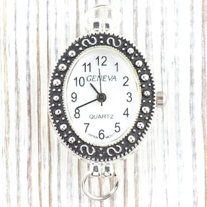 May include: A silver-toned Geneva quartz watch with a white face and black numerals. The oval watch has a decorative bezel and a small ring at the bottom. The watch is approximately 1.5 inches in length.
