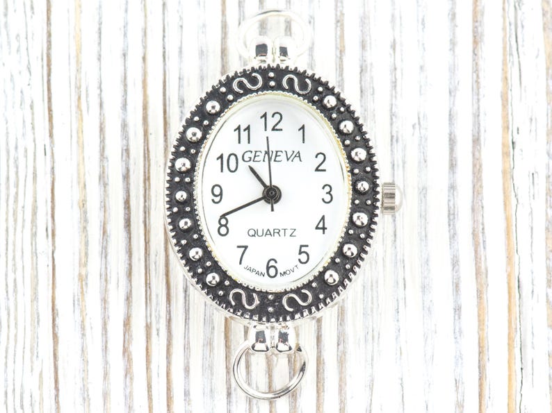 May include: A silver-toned Geneva quartz watch with a white face and black numerals. The oval watch has a decorative bezel and a small ring at the bottom. The watch is approximately 1.5 inches in length.