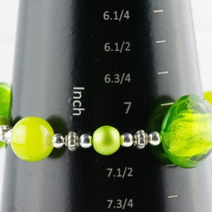 Puede incluir: Un brazalete de cuentas de vidrio verde con cuentas espaciadoras plateadas. El brazalete se mide en una regla que muestra que el brazalete mide 7 pulgadas de largo.