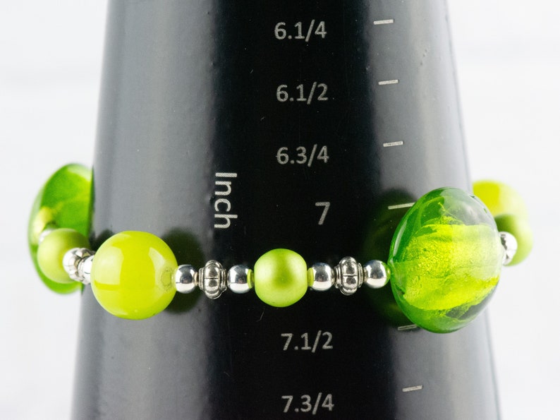 Puede incluir: Un brazalete de cuentas de vidrio verde con cuentas espaciadoras plateadas. El brazalete se mide en una regla que muestra que el brazalete mide 7 pulgadas de largo.