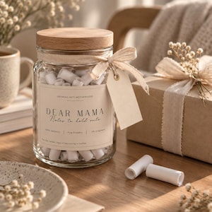 Könnte beinhalten: Ein durchsichtiges Glasgefäß mit einem hellen Holzdeckel, gefüllt mit kleinen weißen Papierrollen. Das Etikett trägt die Aufschrift DEAR MAMA Notes to hold onto, mit einem beigen Band um den Hals und einem leeren rechteckigen Anhänger.