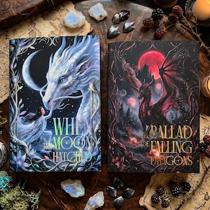 May include: Two fantasy books with intricate covers. One features a white dragon and the title "When the Moon Hatched." The other displays a red dragon and the title "The Ballad of Falling Dragons." The books are surrounded by decorative elements.