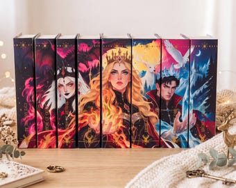 Puede incluir: Un conjunto de seis libros de tapa dura dispuestos uno al lado del otro, con lomos ilustrados vibrantes que muestran personajes de fantasía, incluyendo una reina con fuego, un hombre con hielo y un ciervo blanco.