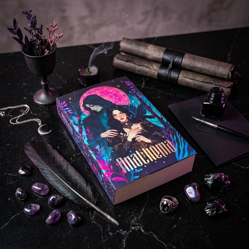 May include: A hardcover book titled "Anathema" with a fantasy illustration of a couple under a pink moon. The image includes a black feather, amethyst stones, a silver necklace, and a small candle with smoke.