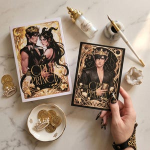 Pode incluir: Duas cópias do livro "The Cruel Prince", uma em capa dura e outra em cartão, com detalhes dourados. Um prato pequeno contém moedas de ouro e uma coroa. Uma garrafa decorativa, caneta e geodo também estão presentes.