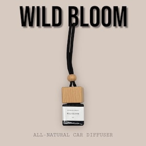 May include: A black car diffuser with a wooden cap, hanging from a black string. The diffuser bottle has a white label that reads "WILD BLOOM" and "ALL-NATURAL CAR DIFFUSER".
