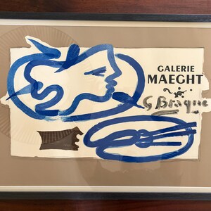May include: Framed art print featuring a blue abstract design of a face and other shapes. The print includes the text "GALERIE MAEGHT" and the signature "G. Braque". The artwork is framed in a black frame.