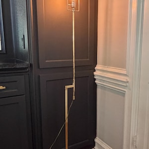 May include: A tall, slender floor lamp with a polished brass finish, featuring a square base, a vertical stem with an adjustable joint, and a rectangular frame at the top holding a single glowing Edison-style light bulb with a visible filament.