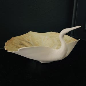 May include: A white ceramic bowl in the shape of a swan, with a long neck and beak. The bowl has a ruffled edge and a light yellow interior. The bowl is a decorative piece.