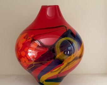 May include: A bulbous glass vase with a vibrant red base color, featuring swirling abstract patterns in shades of deep blue, bright yellow, and orange. The surface is smooth and glossy, with a narrow neck and a wide, rounded body shape.