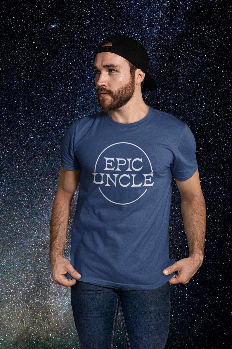 Puede incluir: Un hombre con una camiseta azul con un c&iacute;rculo blanco y el texto "EPIC UNCLE" en su interior. Lleva una gorra de b&eacute;isbol negra y pantalones vaqueros azules. El fondo es azul oscuro con estrellas blancas.
