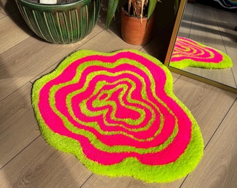 Puede incluir: Una alfombra de forma orgánica irregular con un patrón vibrante y de alto contraste de líneas onduladas concéntricas en colores alternos rosa neón y verde lima. El tejido tiene una superficie texturizada, afelpada y suave.