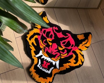Puede incluir: Una alfombra tufting con forma de cabeza de tigre que presenta una paleta de colores vibrantes en negro, naranja brillante, rosa intenso y blanco. El diseño muestra un tigre gruñendo con los dientes al descubierto en hilo texturizado.