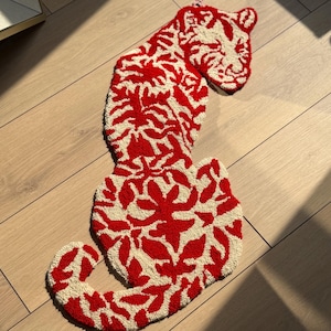 May include: A red and cream-coloured rug shaped like a tiger. The rug has a repeating floral pattern in red on a cream background. The tiger's head and body are clearly defined, creating a striking design.