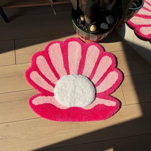Può includere: Un tappeto a forma di conchiglia, rosa e bianco. Il tappeto presenta un bordo esterno fucsia, segmenti rosa più chiari e un cerchio centrale bianco. È posizionato su un pavimento in legno chiaro. Il design evoca un tema costiero.