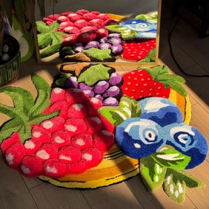 May include: A colorful, tufted rug in the shape of various fruits, including a large red raspberry, purple grapes, and blueberries. The rug has a yellow border and green leaves. The design is reflected in a mirror.