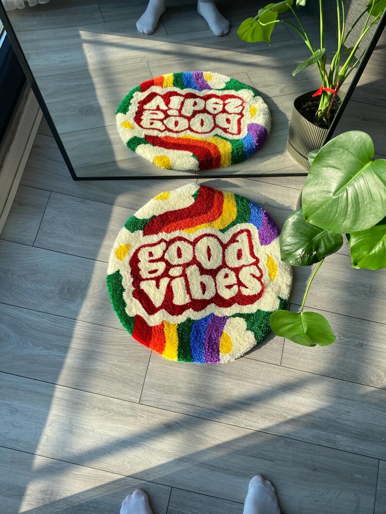 May include: Two round, colorful, fluffy rugs with the words "Good Vibes" and "Good Vibes" in red and white. The rugs have a rainbow border and white daisy-like shapes.