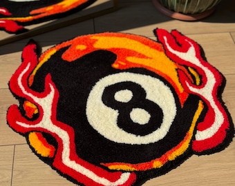 Puede incluir: Una alfombra circular con mechones que presenta un diseño de bola ocho negra en el centro, rodeada de llamas estilizadas en tonos rojo, naranja y blanco. La alfombra tiene una superficie texturizada y afelpada con un borde exterior irregular.