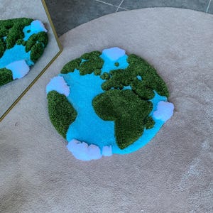 Puede incluir: Una alfombra circular con mechones que presenta un mapa estilizado de la Tierra con masas terrestres de color verde oscuro, un oc&eacute;ano cian brillante y nubes blancas en relieve dispersas por la superficie, reflejadas en un espejo con marco dorado.