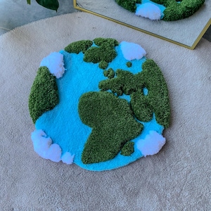 Puede incluir: Una alfombra circular con mechones que presenta un mapa estilizado de la Tierra. El dise&ntilde;o muestra masas terrestres de color verde vibrante, un fondo de oc&eacute;ano azul brillante y secciones blancas en relieve que representan nubes de lana.