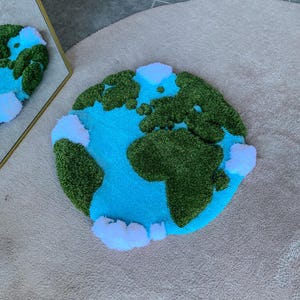 Puede incluir: Una alfombra circular con mechones que presenta un mapa estilizado de la Tierra. El dise&ntilde;o muestra masas terrestres de color verde oscuro sobre un fondo de oc&eacute;ano azul brillante, acentuado con nubes blancas esponjosas alrededor del globo.