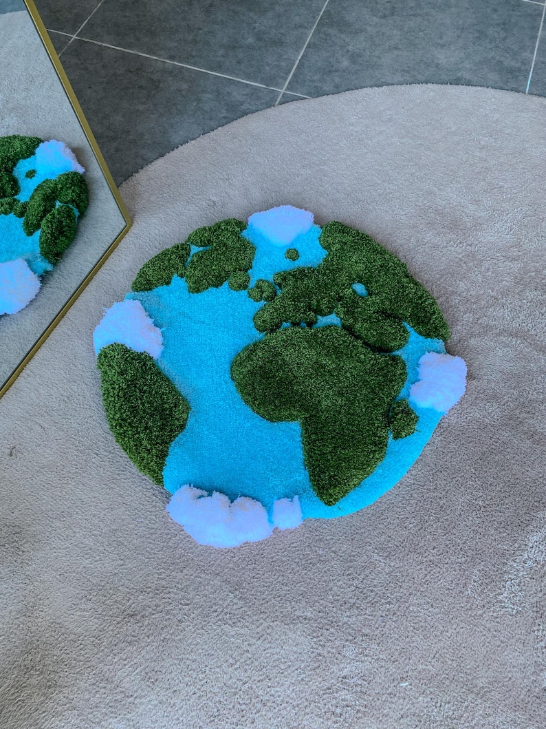 Puede incluir: Una alfombra circular con mechones que presenta un mapa estilizado de la Tierra. El dise&ntilde;o muestra masas terrestres de color verde oscuro sobre un fondo de oc&eacute;ano azul brillante, acentuado con nubes blancas esponjosas alrededor del globo.
