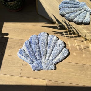 May include: A blue and white seashell-shaped bath mat. The mat is made of a soft, plush material and has a textured surface. The mat is on a wooden floor, with a mirror reflecting the mat. The mat is a decorative item for a bathroom.