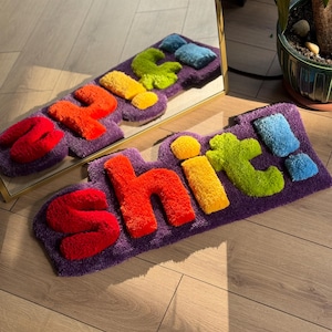 May include: A rectangular purple rug featuring the word "SHIT" in vibrant, raised letters. The letters are in shades of red, orange, yellow, green, and blue, creating a playful design. The rug has a soft, textured finish.