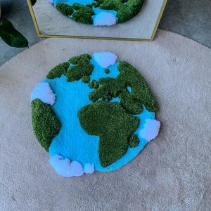 Puede incluir: Una alfombra circular con mechones que presenta un mapa estilizado de la Tierra con masas terrestres verdes texturizadas, un fondo de oc&eacute;ano azul brillante y nubes blancas esponjosas dispersas por el per&iacute;metro, reflejadas en un espejo detr&aacute;s.