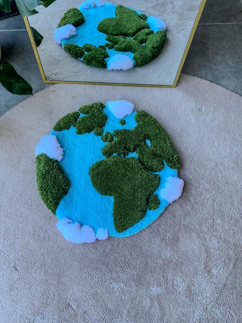 Puede incluir: Una alfombra circular con mechones que presenta un mapa estilizado de la Tierra con masas terrestres verdes texturizadas, un fondo de oc&eacute;ano azul brillante y nubes blancas esponjosas dispersas por el per&iacute;metro, reflejadas en un espejo detr&aacute;s.