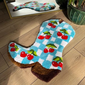 May include: A blue and white checkered bath mat shaped like a cowboy boot with red cherries on it.