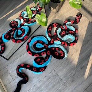 May include: A tufted rug shaped like a coiled snake with a black, bright blue, and red spotted pattern. The rug features a long, winding body with a distinct head and tail, showcasing a high-pile texture throughout the entire surface area.