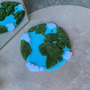 Puede incluir: Una alfombra circular con mechones que presenta un mapa estilizado de la Tierra. El dise&ntilde;o muestra oc&eacute;anos de color azul vibrante, masas terrestres de color verde oscuro texturizado y nubes blancas esponjosas sobre la superficie.