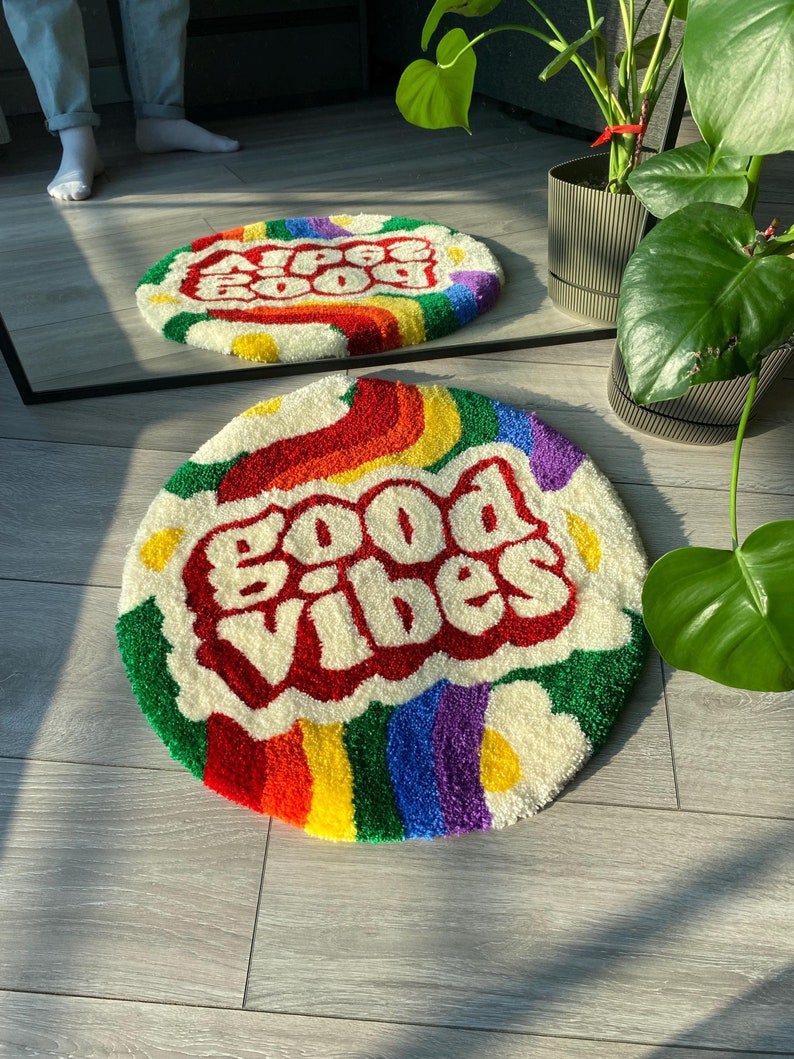 May include: A round, white, fluffy rug with a rainbow border and the words "Good Vibes" in red, outlined in white, in a retro font. The rug is on a wooden floor.