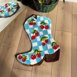 May include: A blue and white checkered boot-shaped rug with red cherries embroidered on it. The rug is on a wooden floor.