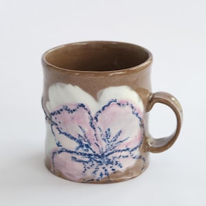 May include: A brown ceramic mug featuring a hand-painted floral design in soft pink and white with dark blue textured detailing. The mug has a cylindrical body, a smooth glossy finish, and a curved handle on the right side of the vessel.