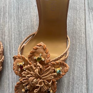 May include: Brown leather sandals with a floral design. The sandals feature a flower made of leather with decorative gemstones in red, green, and clear. Braided straps and a high heel complete the design.