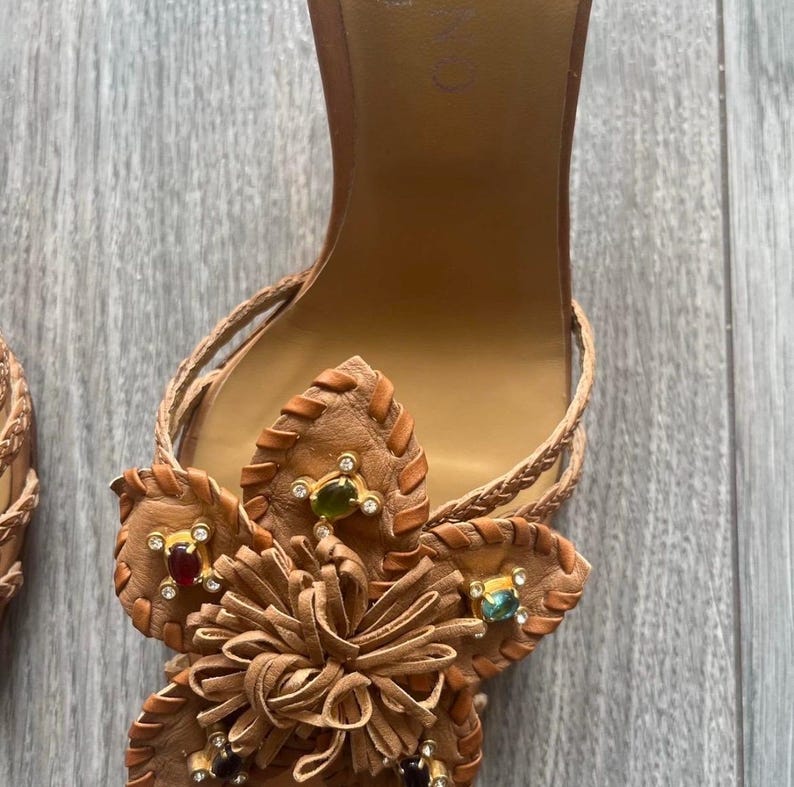 May include: Brown leather sandals with a floral design. The sandals feature a flower made of leather with decorative gemstones in red, green, and clear. Braided straps and a high heel complete the design.