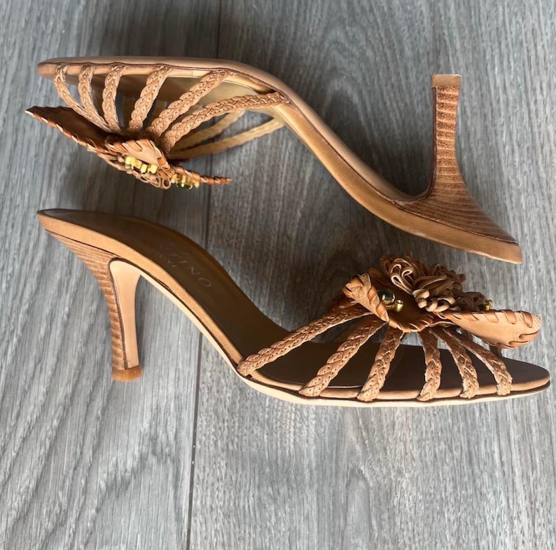 May include: A pair of light brown high-heeled sandals. The sandals have a braided design across the top and a decorative flower with gold and green accents. The heels are approximately 7.6 cm high.