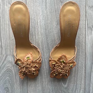 May include: A pair of tan leather Valentino Garavani heeled sandals. Each sandal features a floral embellishment with colourful jewel accents. The sandals have a braided strap design and a high heel. The Valentino Garavani logo is visible on the insole.